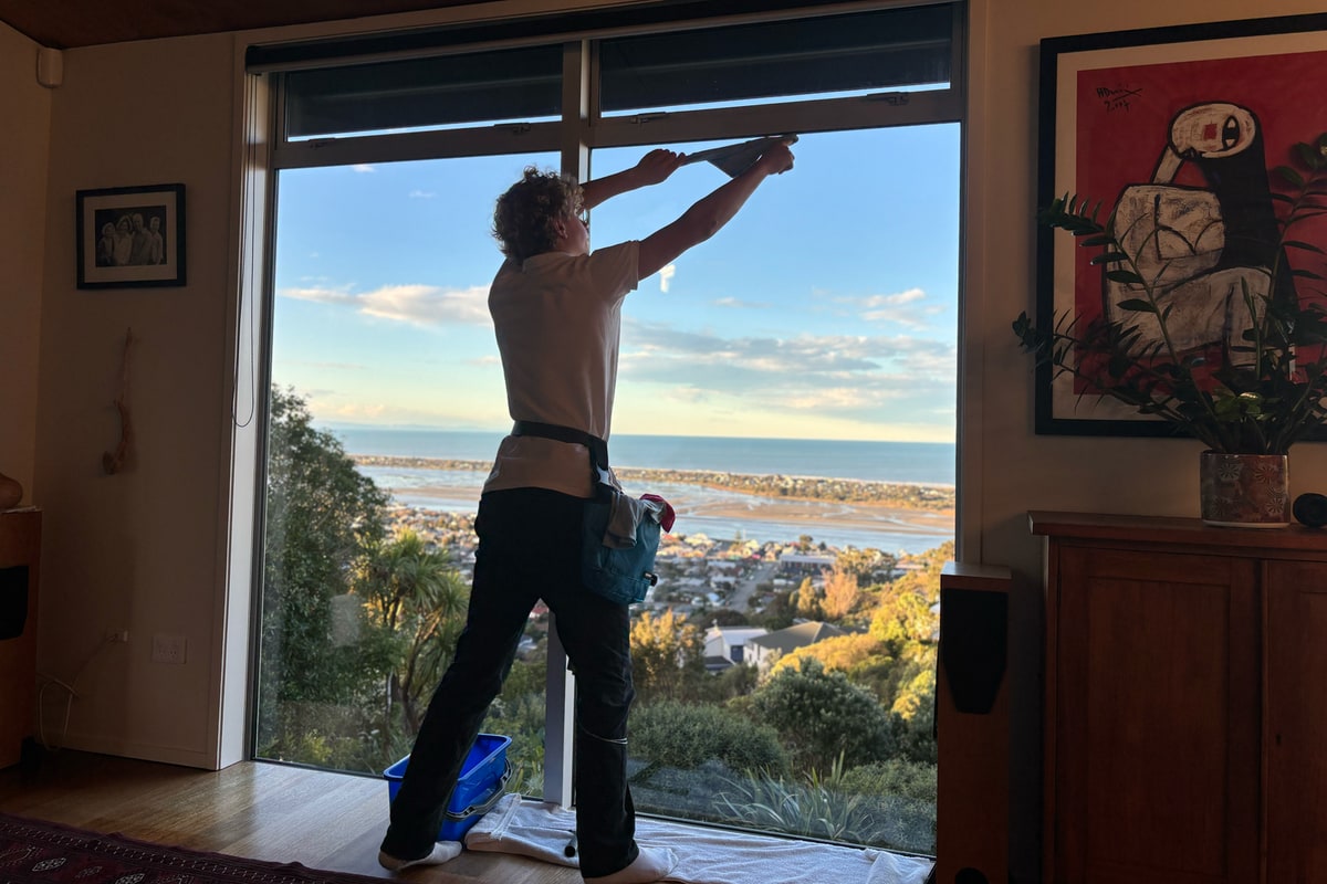 Luca cleaning the inside of a floor-to-ceiling window, drop cloth down, over the estuary